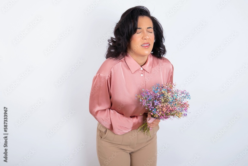 young woman holds big bouquet of nice flowers over white background with hand on stomach because nausea, painful disease feeling unwell. Ache concept. young woman holds big bouquet of nice flowers over white background with hand on stomach because nausea, painful disease feeling unwell. Ache concept.