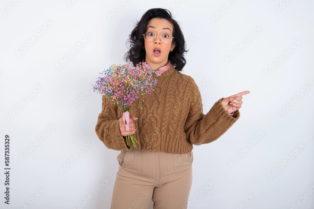 Emotive young woman holds big bouquet of nice flowers over white wall keeps jaw dropped from shock demonstrates amazing promo points right on blank space demonstrates big shopping sale. Advertisement Emotive young woman holds big bouquet of nice flowers over white wall keeps jaw dropped from shock demonstrates amazing promo points right on blank space demonstrates big shopping sale. Advertisement