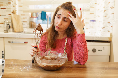 Blonde middle-aged woman with a bowl in front of her in the kitchen is having a problem with her cake. Woman in kitchen with dough beater looking lost in thought.