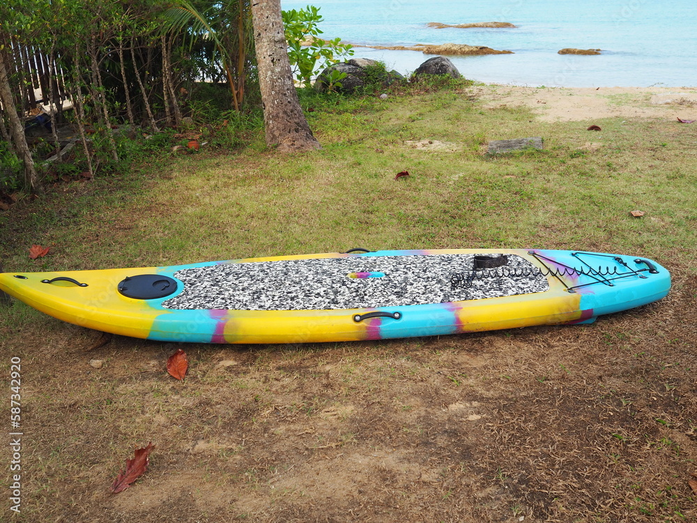 Paddle or SUP Board has big standing area for standing poses and small waves in sea. Is popular and applied sport developed from Surf Board. Yellow-blue stand-up paddle board sits on beach under shade Paddle or SUP Board has big standing area for standing poses and small waves in sea. Is popular and applied sport developed from Surf Board. Yellow-blue stand-up paddle board sits on beach under shade