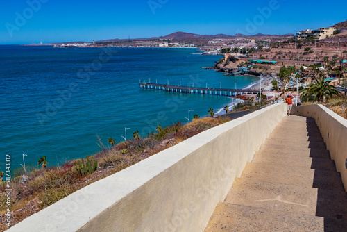 Stairs going up hill to La Ballena viewpoint, coastal coastline, Coromuel beach, pier, sea, and mountains in background against blue sky, woman standing, sunny day La Paz, Baja California Sur Mexico