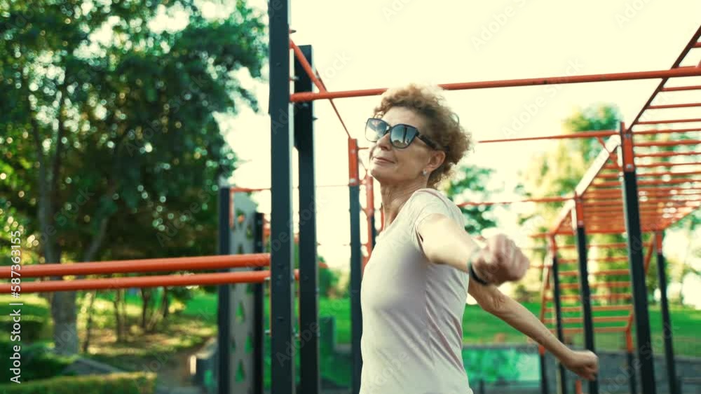 Older female warms up before working out on street fitness site in summer. Elderly woman doing warm-up exercise for arms and upper body belt on outdoor workout area. Retirement healthy way of living. Older female warms up before working out on street fitness site in summer. Elderly woman doing warm-up exercise for arms and upper body belt on outdoor workout area. Retirement healthy way of living.