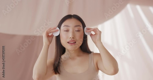 Beautiful Asian woman cleaning face skin. Pretty young female using cotton pad, smoothing skin and removing makeup with micelar water or lotion. Puring, washing, wiping. Skin care, beauty concept. photo
