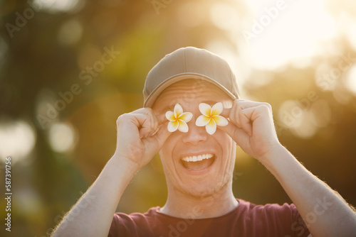 Funny portrait of happy traveler in tropical destination. Man in cap is holding flowers of pumeria in front of his eyes.. photo