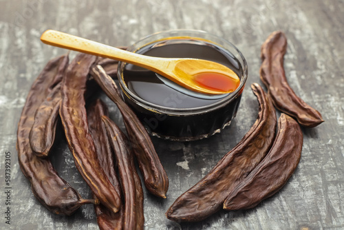 carob molasses in glass bowl and in wooden spoon and carob pods on rustic background, locust bean healthy food, Ceratonia siliqua ( harnup ) photo