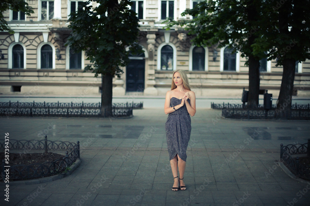 A stylish elegant girl in a dark dress with blond hair stands beautifully in a park alley on a city street, in the evening.