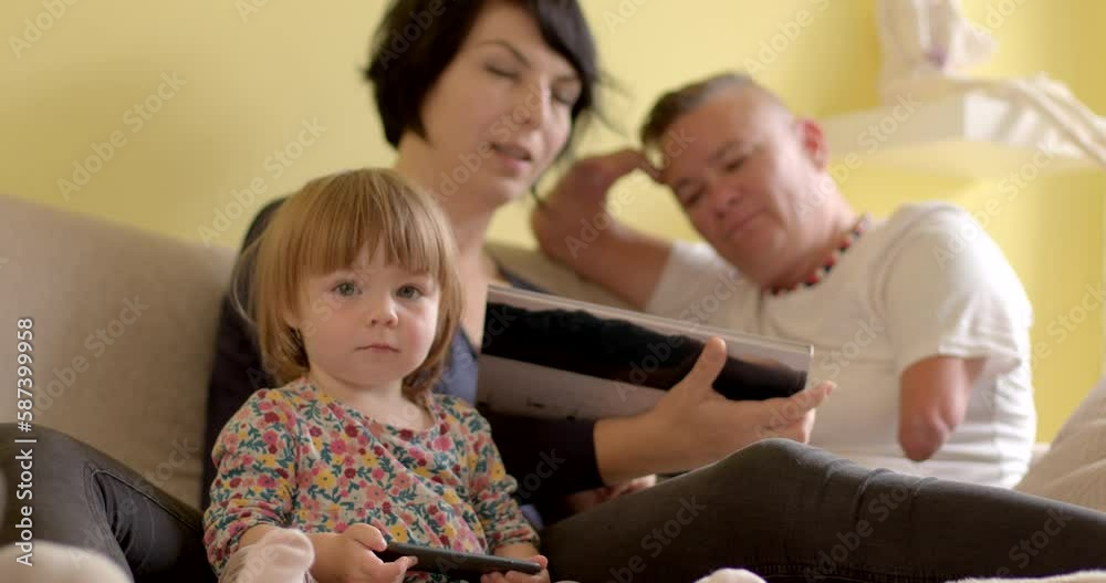 A little girl uses a phone in her hands, she looks into the camera, posing. In the background, the parents are discussing the book. Leisure activities at home on weekends.