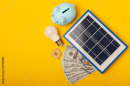 Flat lay composition with solar panel, led lamp, house model, money and piggy bank on yellow background. The concept of saving money and clean energy. Concept of ecology and sustainable development. photo