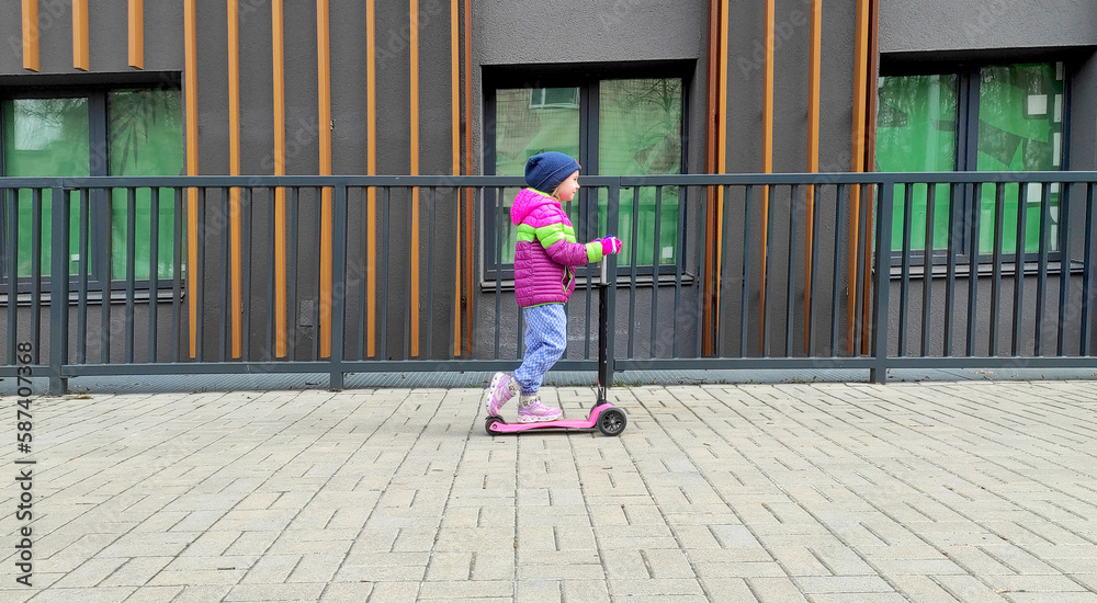 little girl in a multi-colored jacket and hat rides a children's scooter along a brown building with a fence on a city street