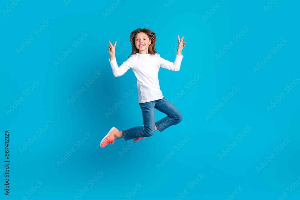 Full length photo of cheerful adorable schoolgirl dressed white shirt jeans flying showing v-sign isolated on blue color background