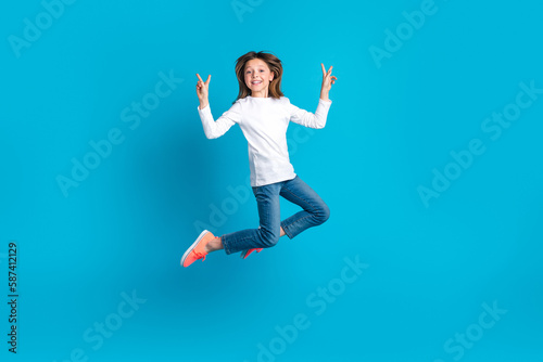 Full length photo of cheerful adorable schoolgirl dressed white shirt jeans flying showing v-sign isolated on blue color background