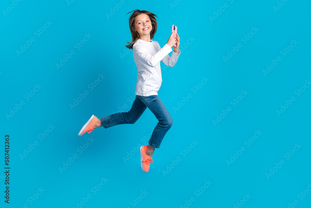 Full length photo of adorable nice schoolgirl dressed white shirt jeans sneakers flying hold smartphone isolated on blue color background