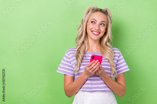 Photo of adorable satisfied girl curly hairdo dressed striped t-shirt hold phone look empty space isolated on green color background