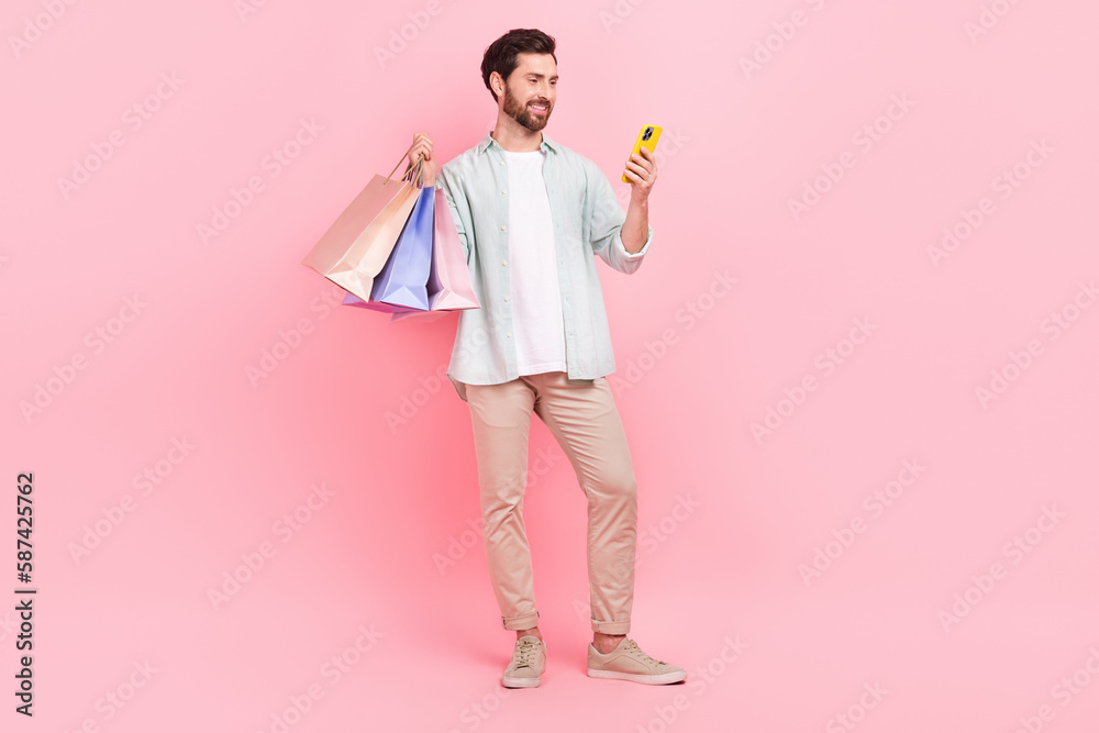 Full length photo of positive cute guy dressed teal shirt holding bargains texting apple samsung gadget isolated pink color background Full length photo of positive cute guy dressed teal shirt holding bargains texting apple samsung gadget isolated pink color background