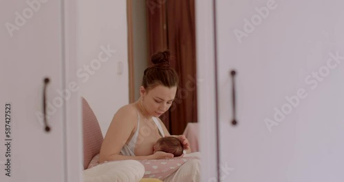 A young mother is sitting on the bed, she is breastfeeding her baby in the room. Feeding the baby in the early stages after childbirth. Reflection in the mirror. Hair in a ponytail.