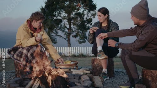 Company of young friends sitting at glampsite on mountain peak, cooking corn on the cob over campfire, having drinks, smiling and having discussion photo