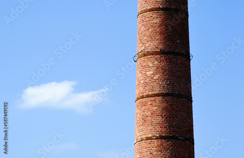 tall tapered large brick chimney stack closeup. rusty metal reinforcing straps. clear blue sky with white cloud. abstract composition. smoke, pollution and environment concept. large diameter cylinder