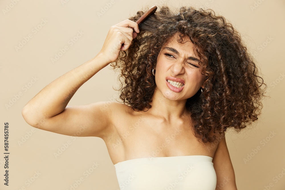 Woman combs curly afro hair with wooden comb, home beauty care styling products, hair loss after covid-19, smile on beige background Woman combs curly afro hair with wooden comb, home beauty care styling products, hair loss after covid-19, smile on beige background