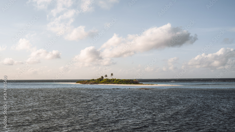 A Maldivian islet features a lush population of green trees with a bit of sand peeking through. Surrounded by the vast blue sea, white clouds in the sky contrast with the overall calming scene