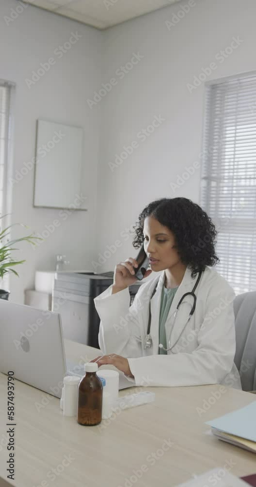 Vertical video of biracial female doctor talking on smartphone and using laptop, slow motion Vertical video of biracial female doctor talking on smartphone and using laptop, slow motion