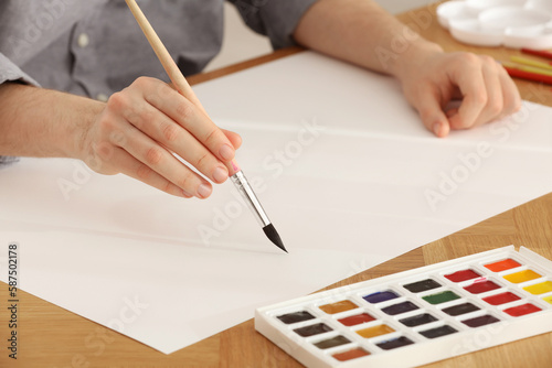 Man painting with watercolor on blank paper at wooden table, closeup