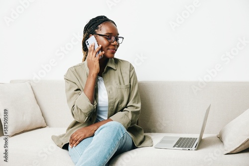 African American woman business freelancer working sitting on sofa at home in laptop and phone, business calls and correspondence happiness smile, home clothes eyeglasses, light interior background.