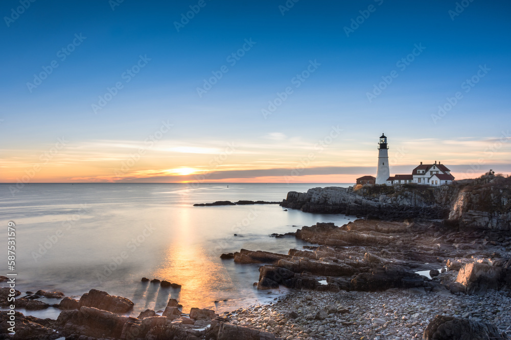 Fototapeta premium Sunrise at Portland Head Lighthouse, Portland, Maine. Looking across the water and rocks towards the lighthouse. Rays gleaming across the water shining on four rocks casting shadows