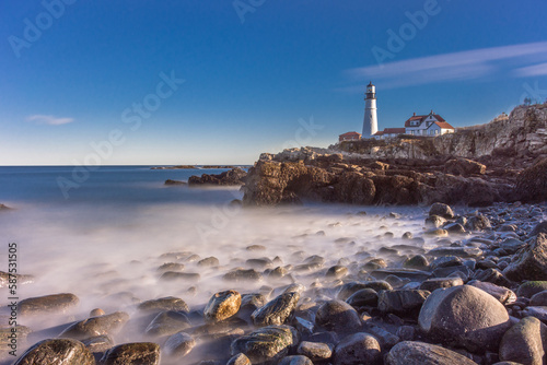 Portland Head Lighthouse, Portland Maine. Lighthouse on cliff with keeper's quarters. Shot from ground level below cliffs. Bright sunshine. Long exposure water misting over rocks in foreground.