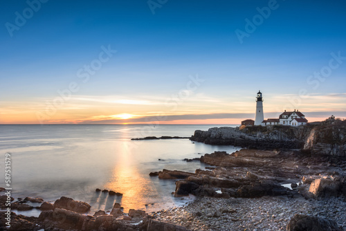 Sunrise at Portland Head Lighthouse, Portland, Maine. Looking across the water and rocks towards the lighthouse. Rays gleaming across the water shining on four rocks casting shadows