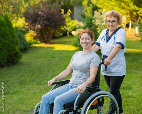 An elderly female nurse walks with a middle-aged woman in a wheelchair in the park. 