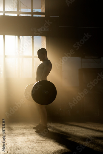 Full shot of a shirtless young athlete man lifting a heavyweight barbell in gym