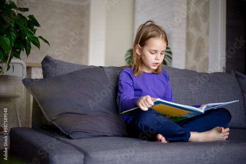 a little blonde girl is reading a big book sitting on the sofa in the living room.Smile and interest in reading.Self-education of children, reading literature at home, interesting leisure