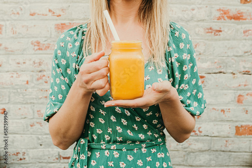 A woman is holding a mango and turmeric vegan smoothie in a glass jar.