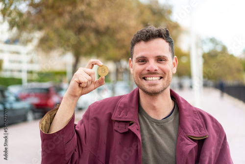 Young caucasian man holding a Bitcoin at outdoors smiling a lot