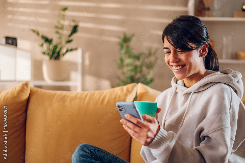 Beautiful woman using smartphone and drinking coffee on the sofa at home Beautiful woman using smartphone and drinking coffee on the sofa at home