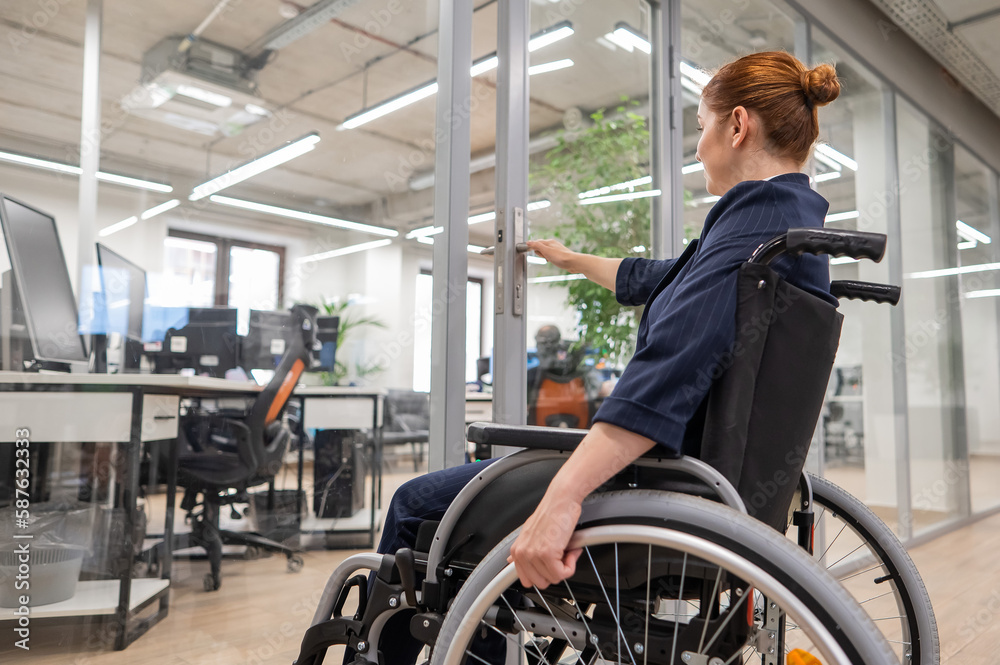 Red-haired caucasian woman in a wheelchair trying to open the door in the office. Red-haired caucasian woman in a wheelchair trying to open the door in the office.