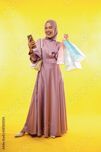 happy Asian muslimah woman stands carrying cell phone with shopping bag on yellow background