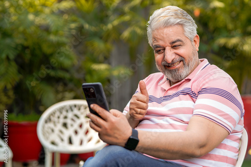 Happy senior man sitting at garden and using smartphone.
