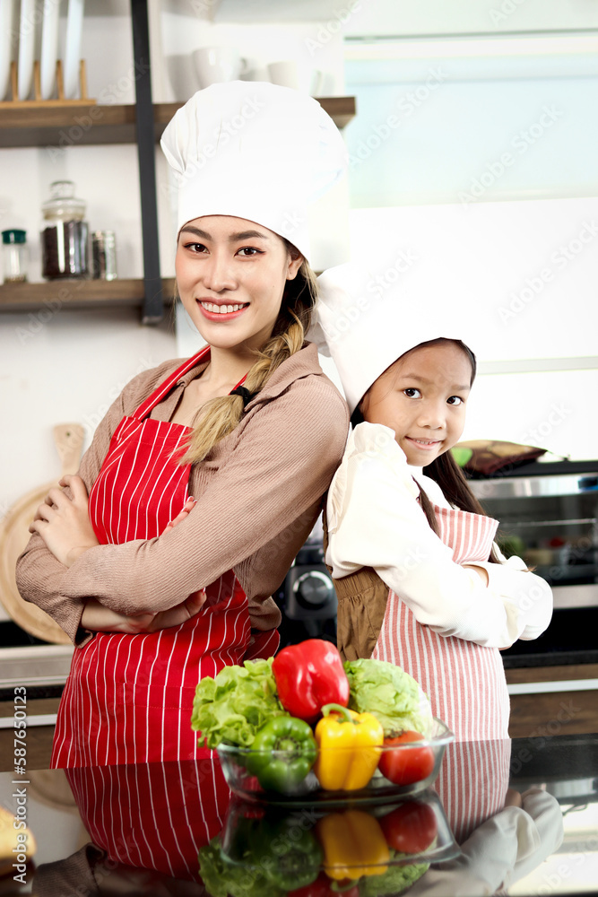 Portrait of happy smiling Asian mother daughter wear apron chef hat standing with arms crossed at kitchen with fresh vegetables salad food ingredient, cute chef family prepare cooking healthy food. Portrait of happy smiling Asian mother daughter wear apron chef hat standing with arms crossed at kitchen with fresh vegetables salad food ingredient, cute chef family prepare cooking healthy food.