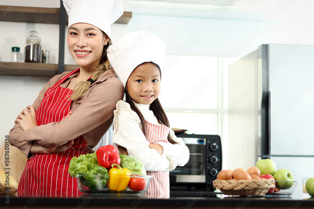 Portrait of happy smiling Asian mother daughter wear apron chef hat standing with arms crossed at kitchen with fresh vegetables salad food ingredient, cute chef family prepare cooking healthy food. Portrait of happy smiling Asian mother daughter wear apron chef hat standing with arms crossed at kitchen with fresh vegetables salad food ingredient, cute chef family prepare cooking healthy food.