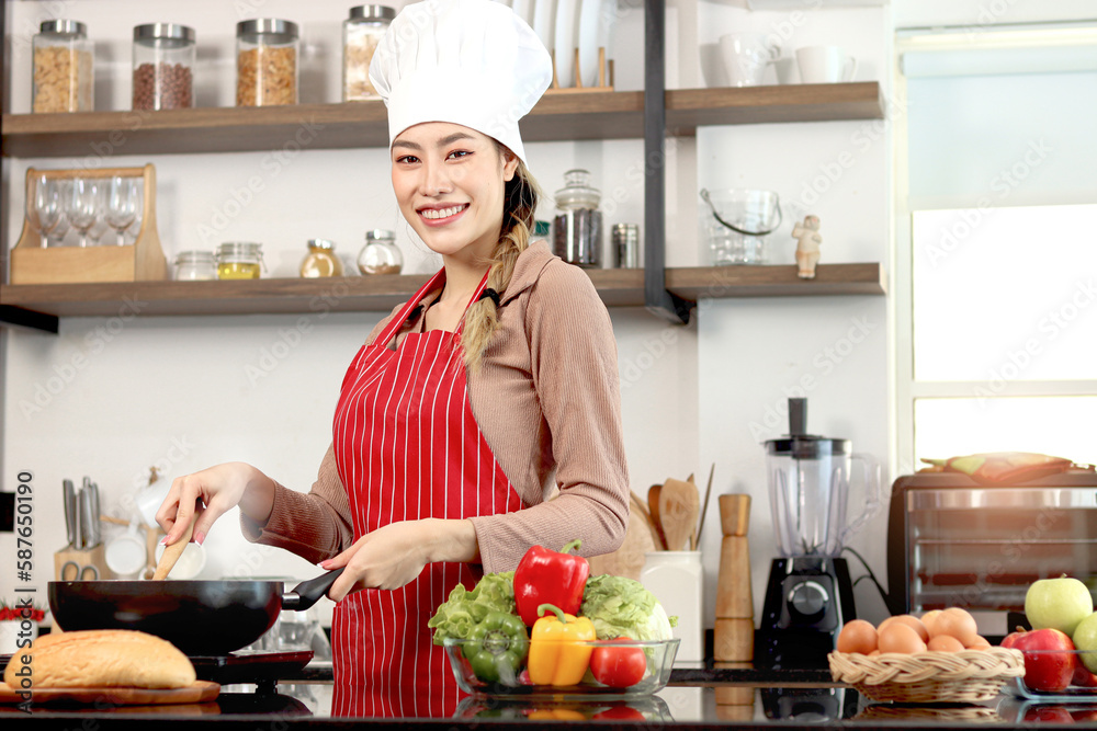 Happy smiling beautiful Asian woman wearing apron and chef hat, holding turner for cooking tasty fried food meal with pan at kitchen with fresh vegetable ingredient, female chef cooking healthy food. Happy smiling beautiful Asian woman wearing apron and chef hat, holding turner for cooking tasty fried food meal with pan at kitchen with fresh vegetable ingredient, female chef cooking healthy food.