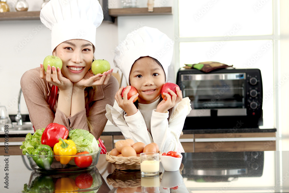 Portrait of happy Asian mother and daughter with apron chef hat holding apples during cooking healthy meal at kitchen, lovely family chef cooking food at home, mom and child spending time together. Portrait of happy Asian mother and daughter with apron chef hat holding apples during cooking healthy meal at kitchen, lovely family chef cooking food at home, mom and child spending time together.