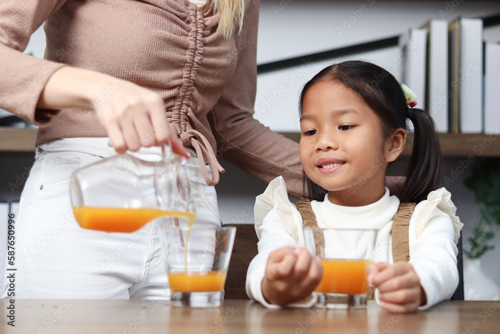 Asian happy family spending time together, mother pouring fresh orange juice from jug for her little cute daughter sitting in living room at home, young parent mom taking care kid to get healthy diet. Asian happy family spending time together, mother pouring fresh orange juice from jug for her little cute daughter sitting in living room at home, young parent mom taking care kid to get healthy diet.