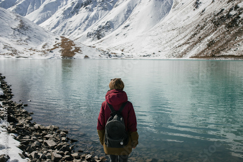A medium shot photo of a woman standing alone with a backpack and looking at a high mountain lake