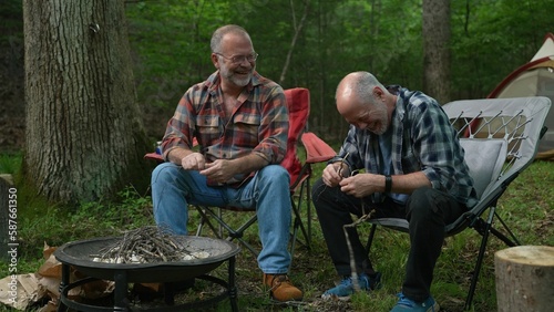 Two gay men building a campfire in a forest talking and laughing with tent in background.