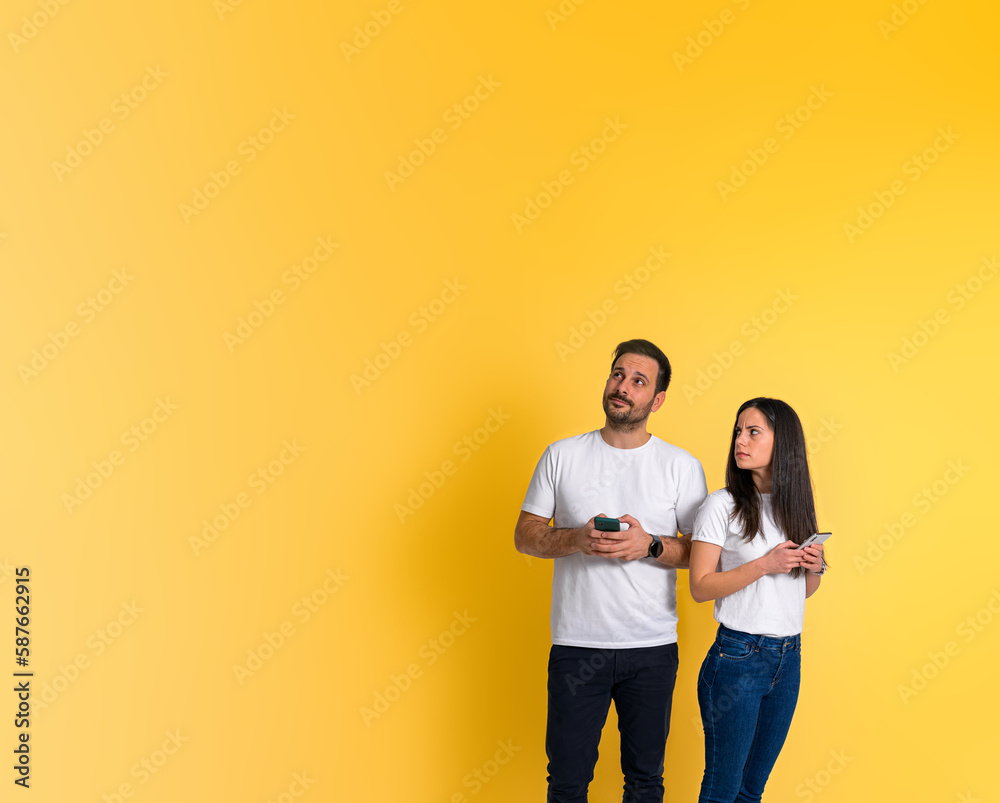 Angry young adult girlfriend staring at possessive spying boyfriend looking away while chatting on smart phone isolated against yellow background Angry young adult girlfriend staring at possessive spying boyfriend looking away while chatting on smart phone isolated against yellow background