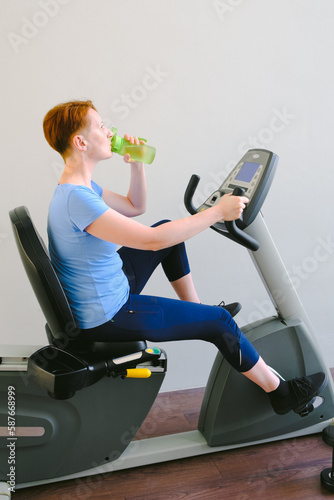 Woman sitting on exercise bike in the gym and drinking water from a sports bottle. Middle-aged woman in a blue t-shirt goes in for sports. The concept of a healthy lifestyle, weight loss. photo