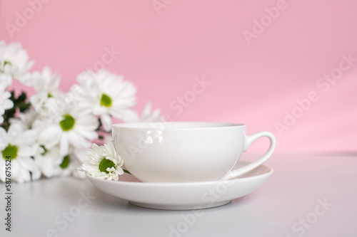 A white cup with a plate stands on a pink background with white chrysanthemum flowers
