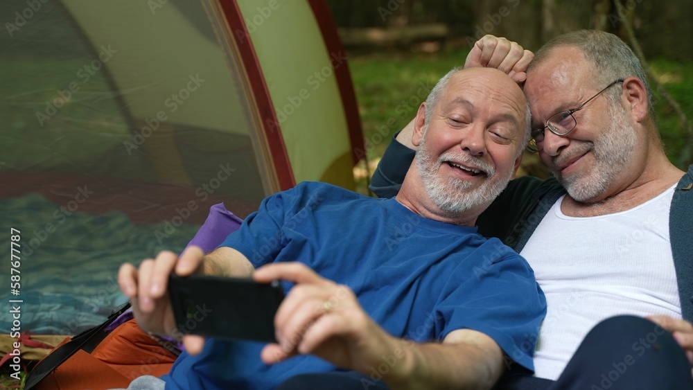 Closeup of two gay men in front of tent with pride flag at campsite taking selfie with phone.