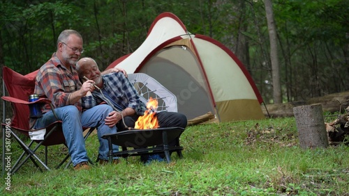 Two gay men in embracing in front of campfire camping in forest with tent.